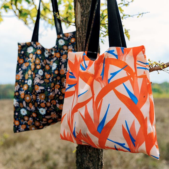 Zwei Tragetaschen mit unterschiedlichen Mustern hängen an einem Baum. Eine Tasche hat ein florales Muster, die andere ein abstraktes, farbenfrohes Design.