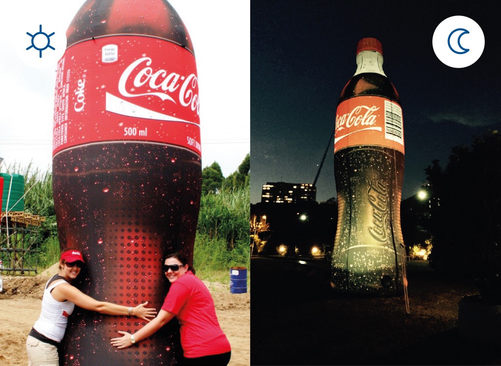 Zwei Frauen umarmen eine große Coca-Cola-Flasche, die bei Tag und Nacht beleuchtet ist.