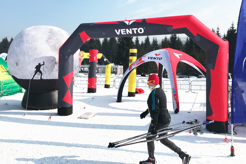 Ein Skifahrer mit Skistöcken geht unter einem aufblasbaren Tor mit dem Logo 'VENTO' hindurch, umgeben von Schnee und weiteren aufblasbaren Elementen.