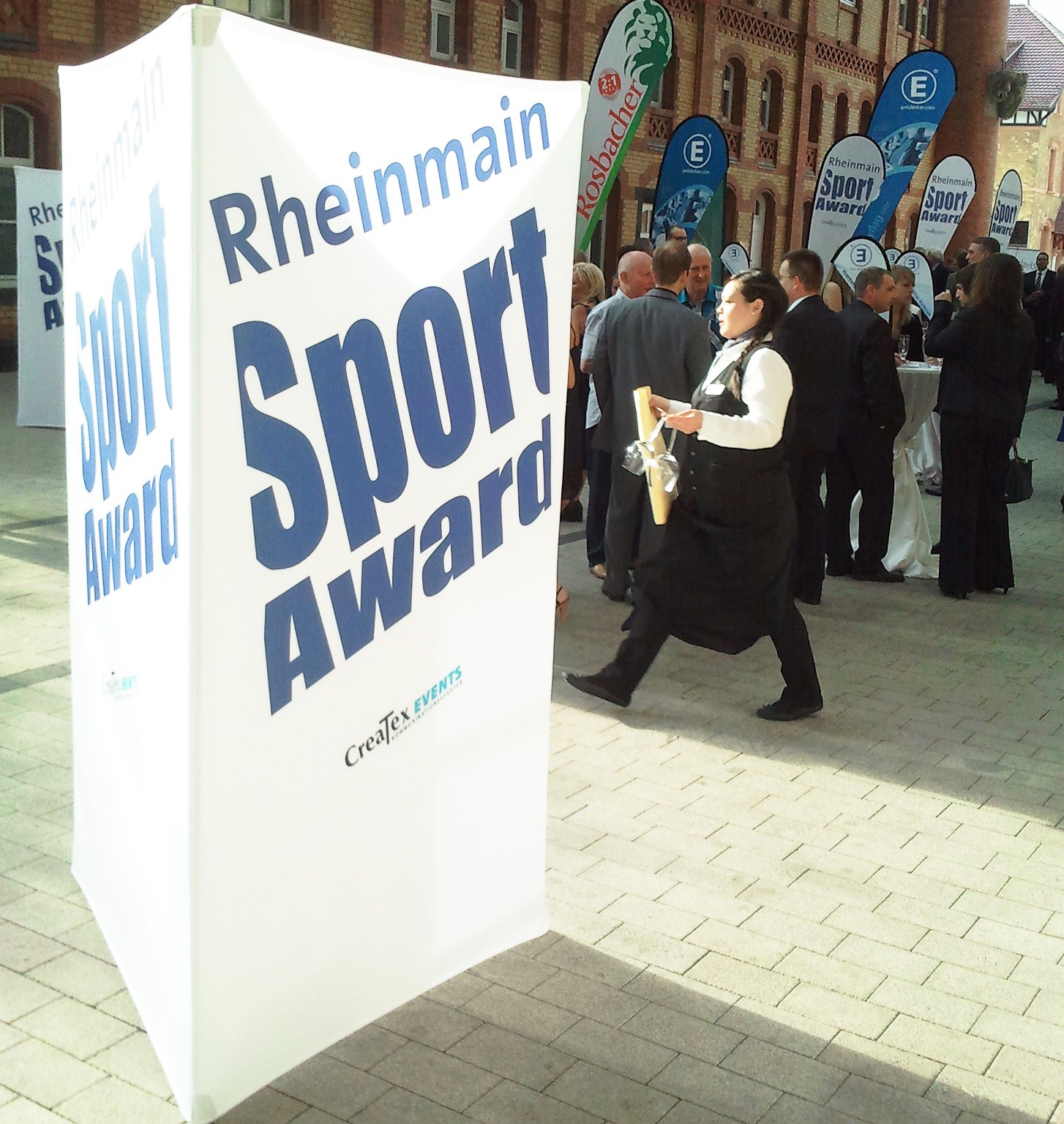 Werbesäule mit der Aufschrift 'Rheinmain Sport Award' auf einer Veranstaltung, umgeben von Gästen in formeller Kleidung.