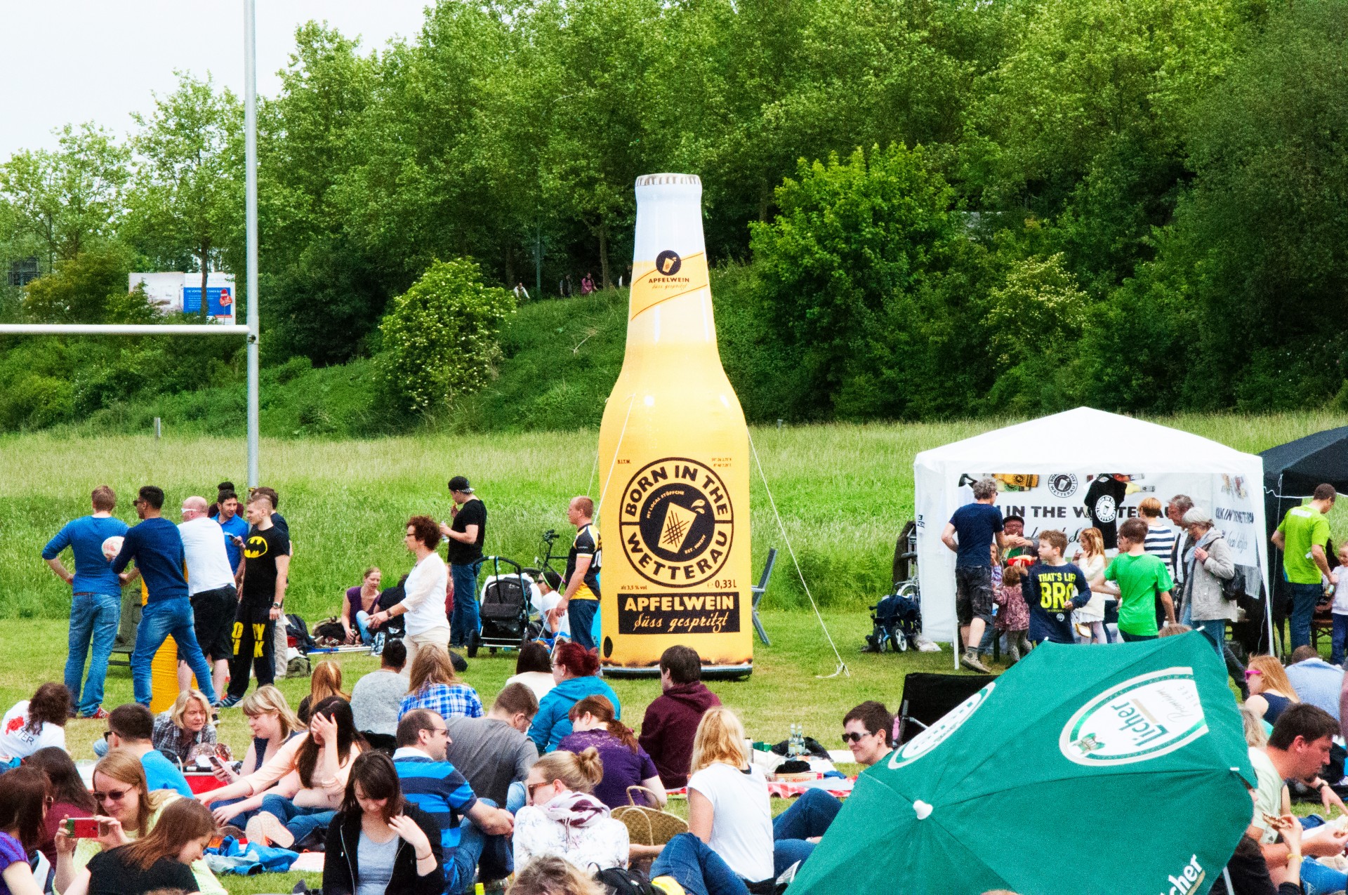 Auf einem Festivalgelände steht eine große aufblasbare Flasche mit der Aufschrift 'Born In The Wetterau'. Menschen sitzen auf dem Gras und genießen die Veranstaltung.