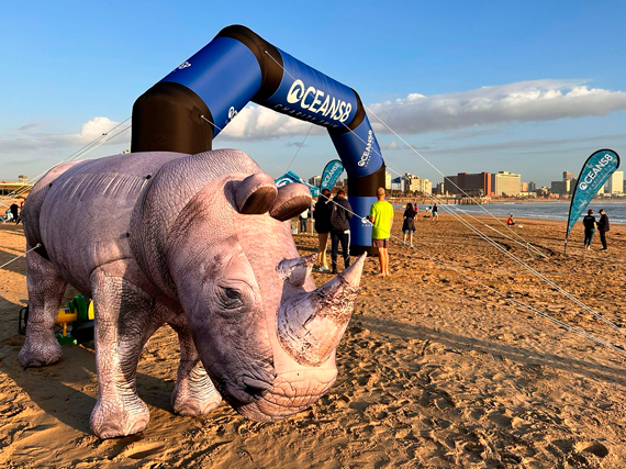 Ein großer, aufblasbarer Nashorn steht am Strand unter einem Bogen mit der Aufschrift 'OCEANSB'. Im Hintergrund sind Menschen und Gebäude sichtbar.