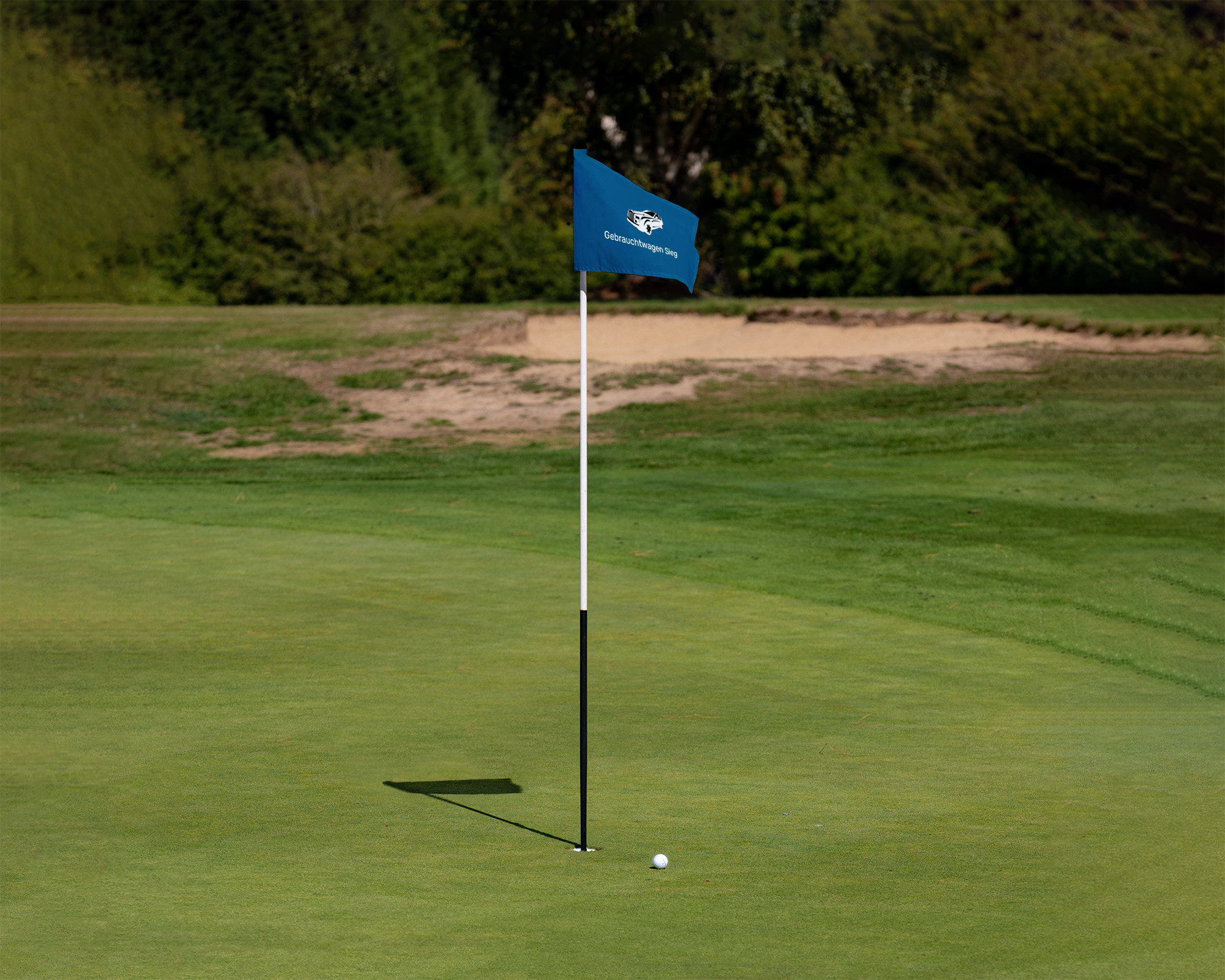 Ein Golfball liegt nahe einer Fahne auf einem Golfplatz mit grünem Rasen im Hintergrund.