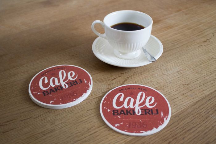 Zwei runde Untersetzer mit der Aufschrift 'Cafe Bakudrij 1976' neben einer Tasse Kaffee auf einem Holztisch.