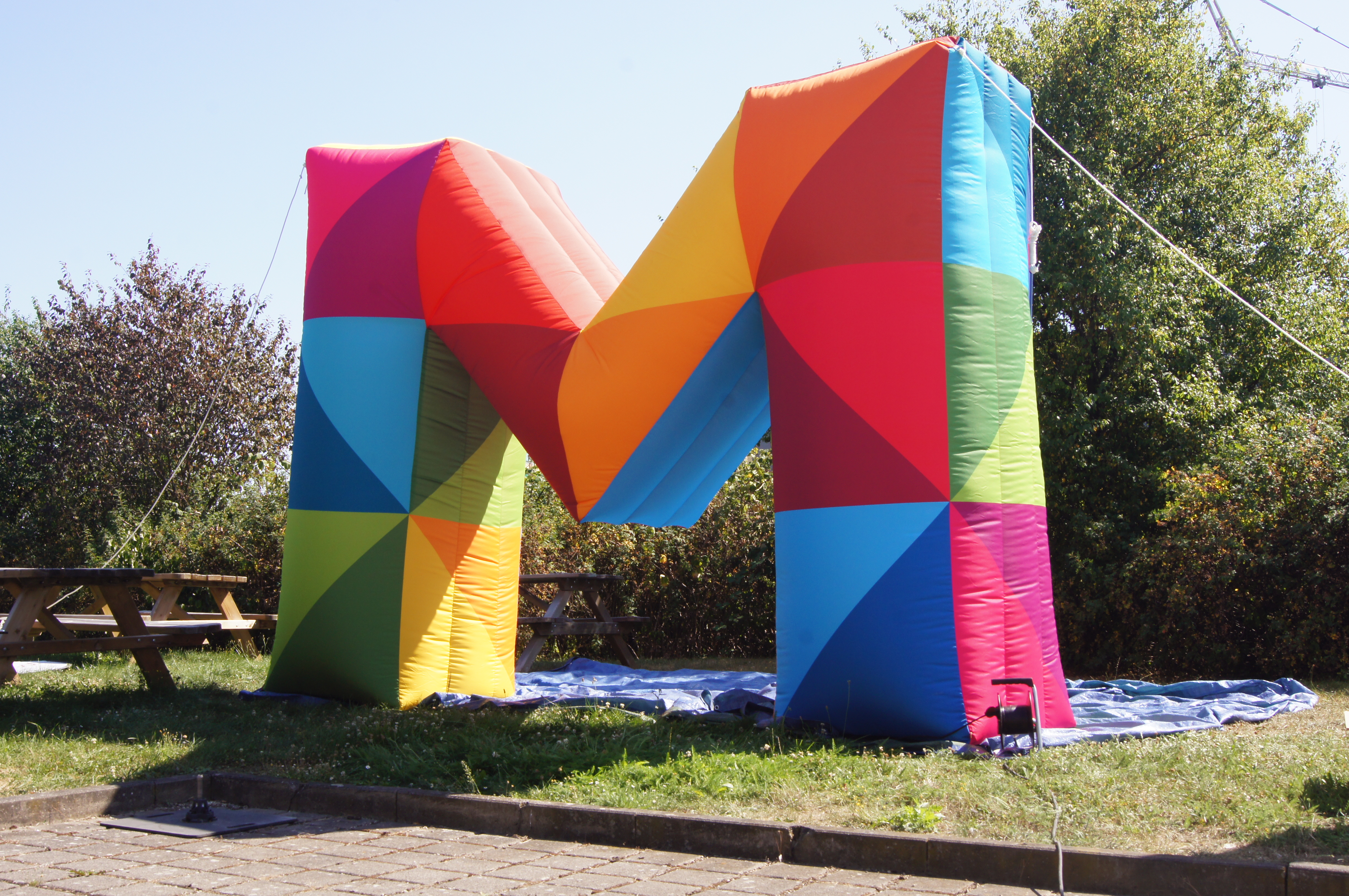 Buntes, aufblasbares M in geometrischen Mustern, auf einer Wiese mit Bäumen im Hintergrund.