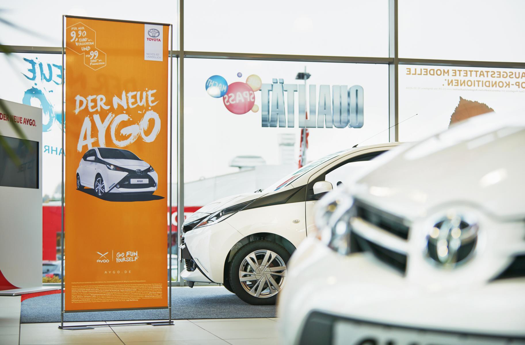 Roll-Up Easymag Stand von Toyota mit dem Slogan 'Der neue Aygo' in einem Autohaus, im Hintergrund sind Autos sichtbar.