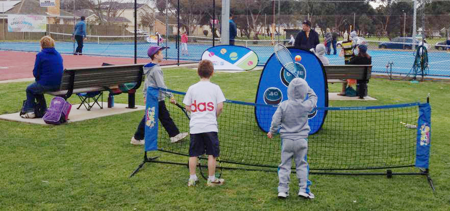 Zwei Kinder spielen mit einer mobilen Tennis Zielscheibe namens Quickboard auf einem Tennisplatz, während Erwachsene zuschauen.