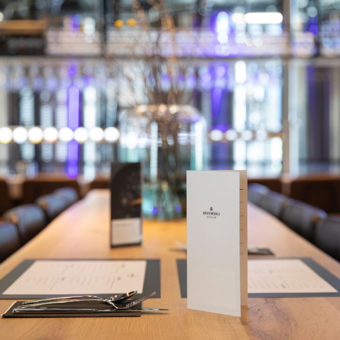 Zwei Speisekarten auf einem Tisch in einem Gastronomiebetrieb, mit Besteck und einem Glas im Hintergrund.
