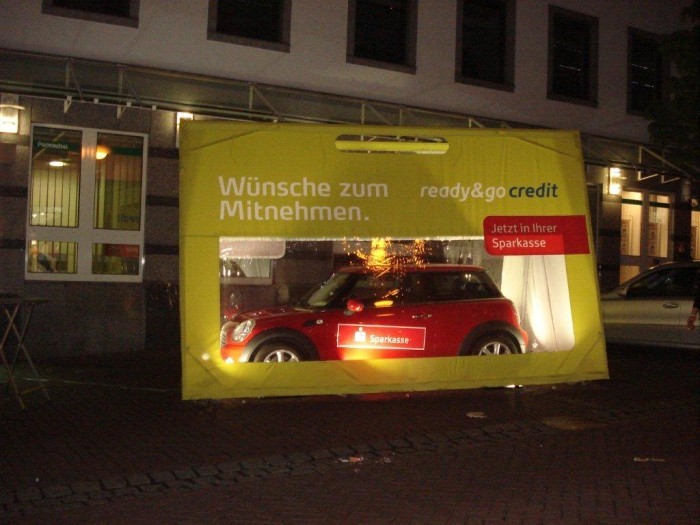 Eine Autotasche mit einem Mini Cooper darin, beleuchtet bei Nacht. Auf der Tasche steht 'Wünsche zum Mitnehmen. ready&go credit. Jetzt in Ihrer Sparkasse'.