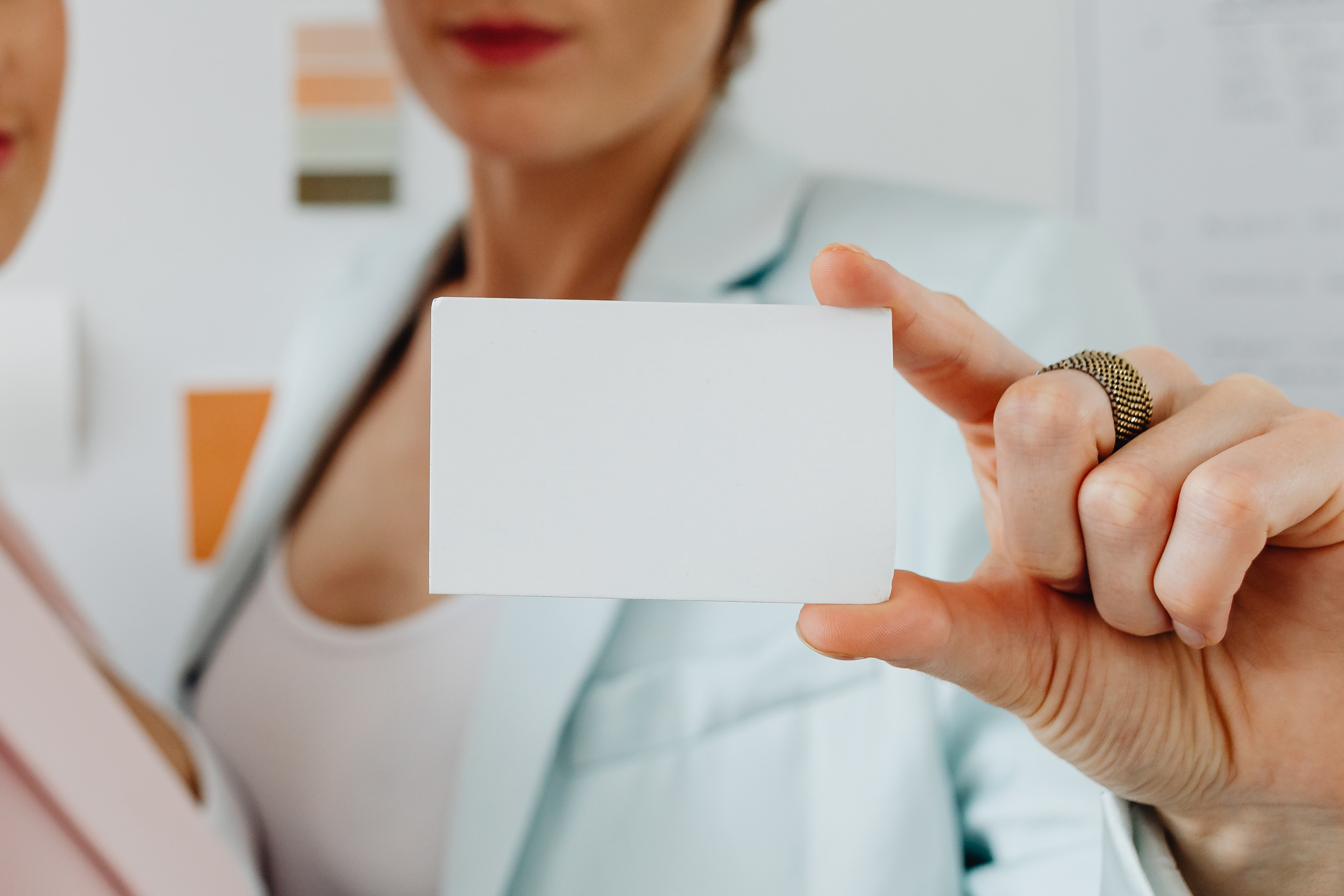 Zwei Frauen stehen vor einer Wand mit Farbmustern und halten eine leere Karte in der Hand. Eine Frau trägt einen blauen Blazer, die andere einen rosa Blazer.