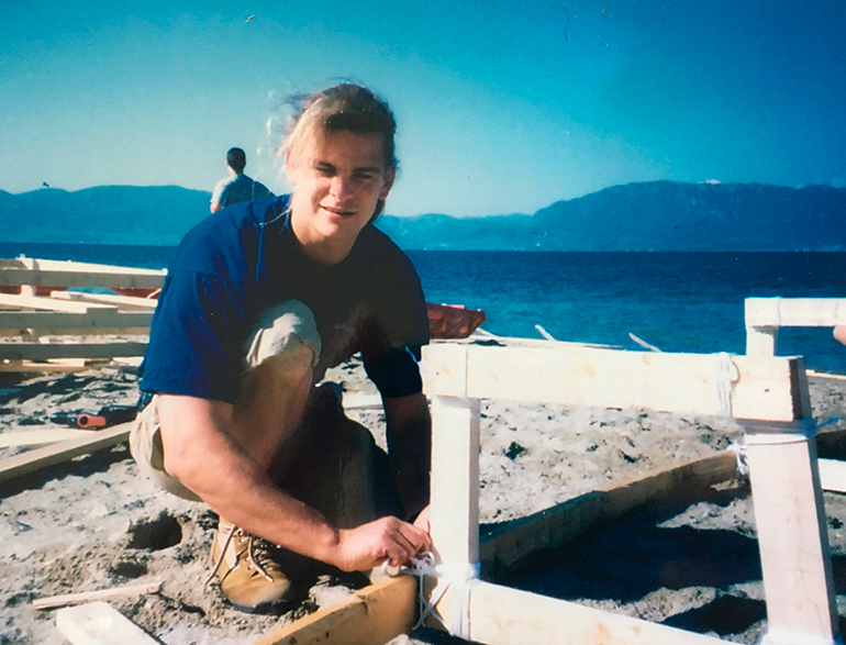 Ein junger Mann mit langen Haaren kniet am Strand und arbeitet an einem Holzrahmen für den Bootsbau.