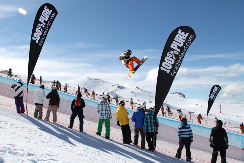 Snowboarder springt über eine Halfpipe, umgeben von Zuschauern und Beachflags mit dem Text '100% PURE'.