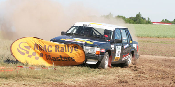 Ein Volvo-Rennwagen fährt durch einen staubigen Feldweg und passiert ein Banner der ADAC Rallye Masters.