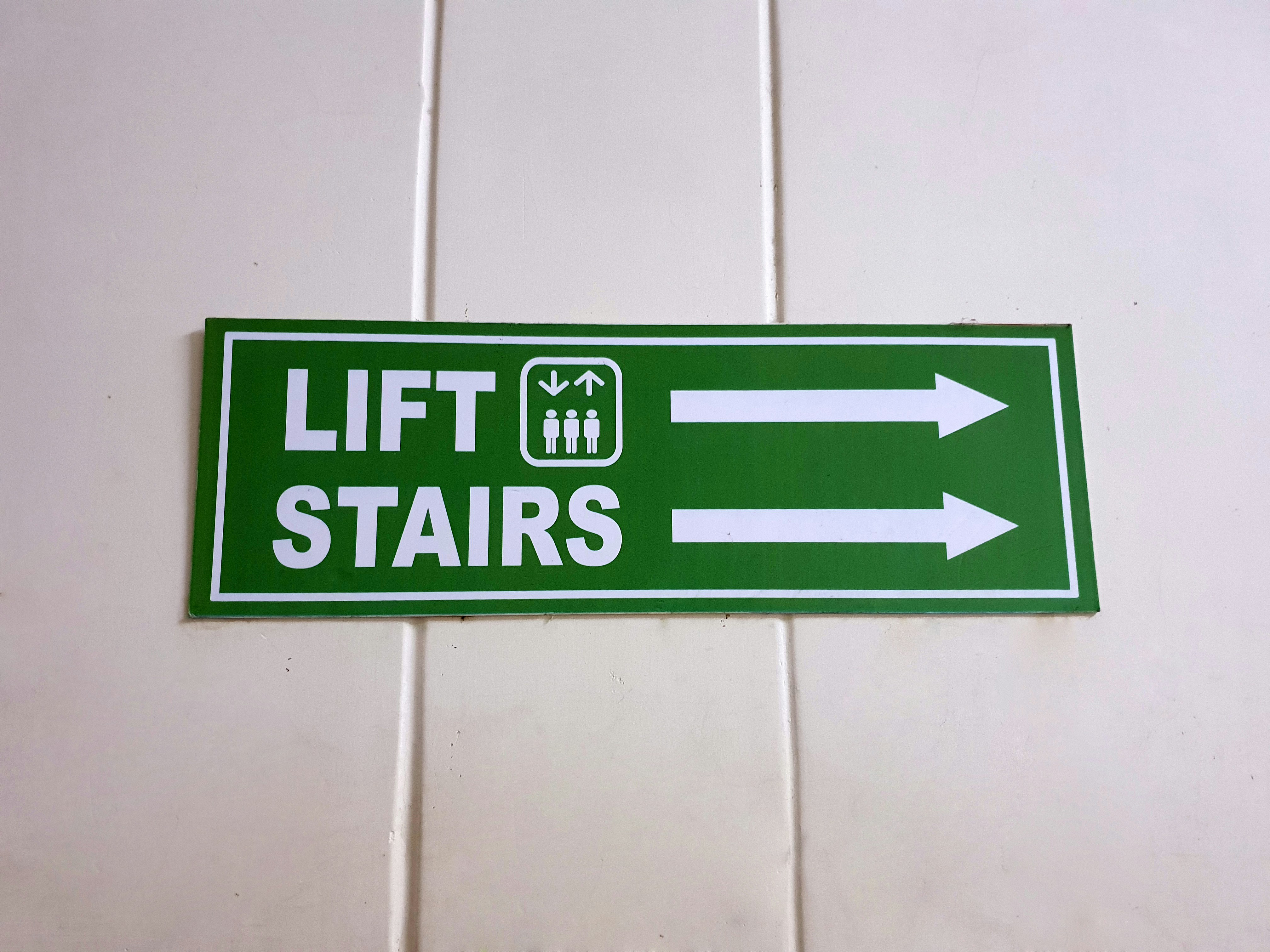 Grünes Werbeschild mit den Aufschriften 'LIFT' und 'STAIRS' sowie Pfeilen nach links und rechts.