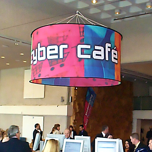 Runder Deckenhänger mit der Aufschrift 'Cyber Café' im Internetcafé, umgeben von Besuchern und Computerarbeitsplätzen.