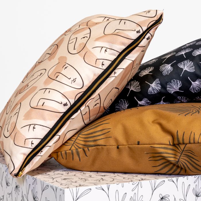 Dekokissen Stapel in verschiedenen Designs: ein Kissen mit Gesichtern, ein schwarzes Kissen mit floralen Mustern und ein braunes Kissen. Alle haben einen Reißverschluss.