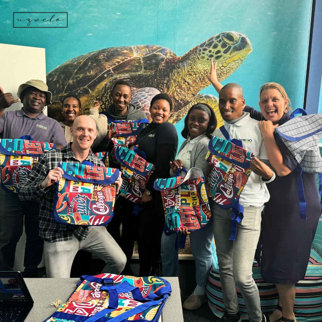 Gruppe von acht Personen, die bunte Taschen mit verschiedenen Designs halten, vor einem Wandbild einer Schildkröte.