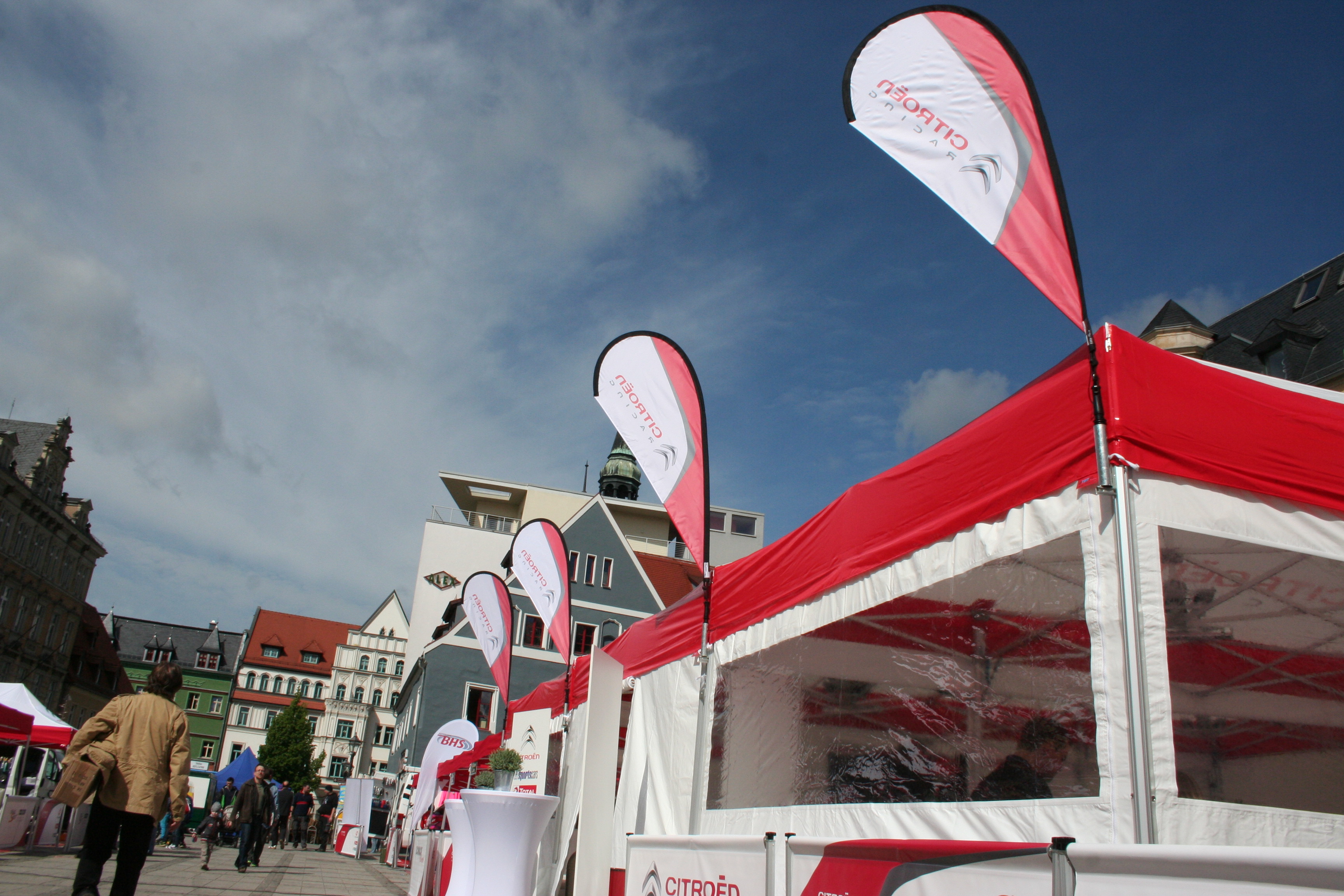 Eventzelte mit roten und weißen Beachflags von ENTDECKER auf einem Platz mit Menschen und Gebäuden im Hintergrund.