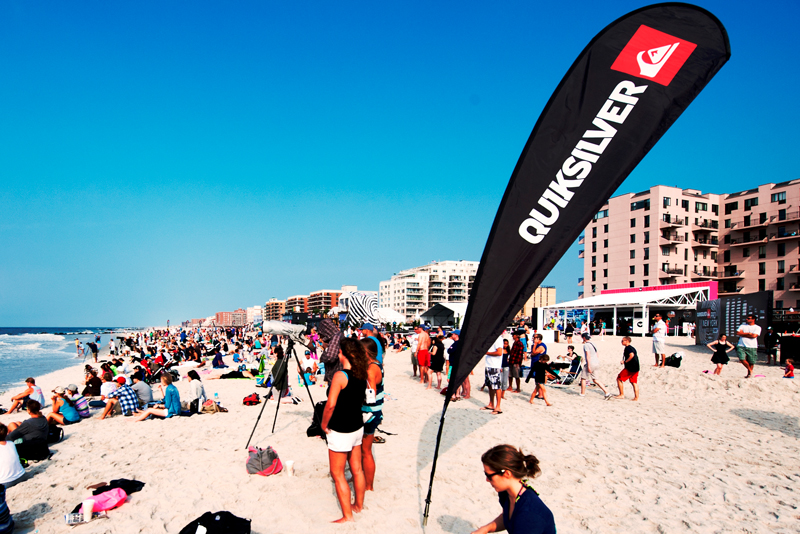 Beachflag von Quiksilver am Strand, umgeben von Menschen und Gebäuden im Hintergrund.