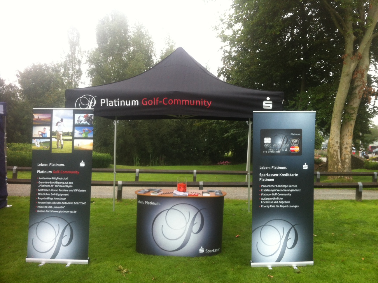 Stand der Platinum Golf-Community mit zwei Roll-Ups und einem Tisch im Freien, umgeben von Bäumen.