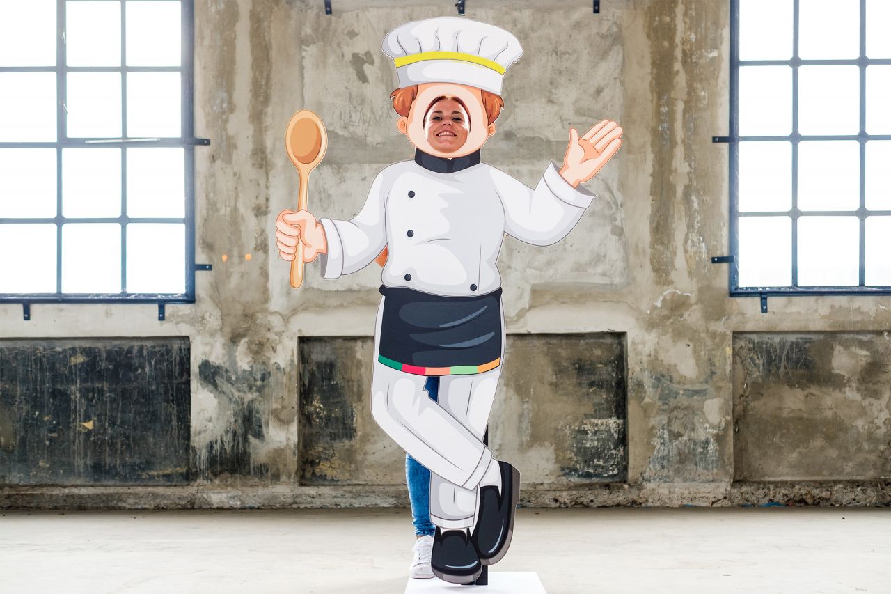 Aufsteller einer Kochfigur mit Kochmütze und Kochlöffel, steht in einem Raum mit großen Fenstern.