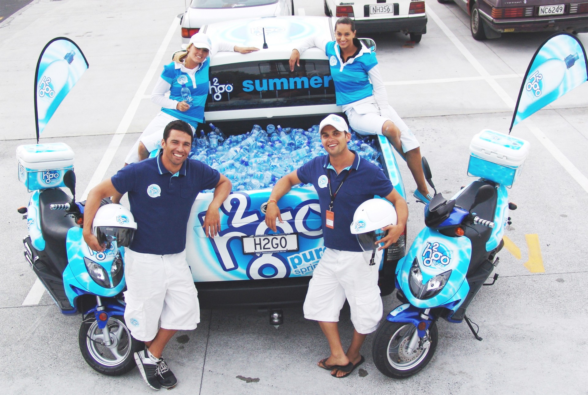 Drei Personen in blauen H2GO-T-Shirts stehen vor einem Fahrzeug mit Wasserflaschen und H2GO-Logo. Zwei Motorräder sind sichtbar.