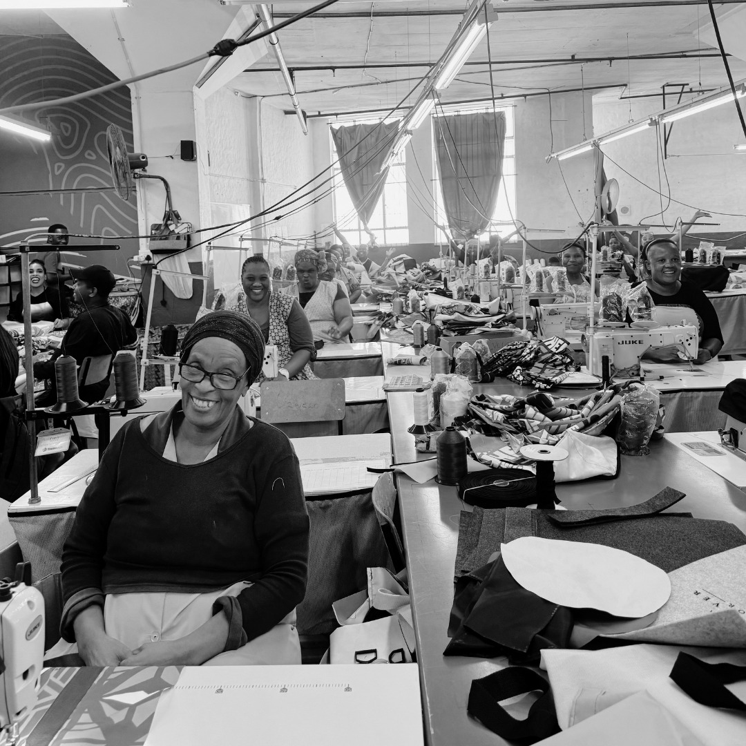 Gruppe von Frauen in einer Nähwerkstatt, die an Nähmaschinen arbeiten und lächeln. Stoffe und Nähmaterialien sind auf den Tischen verteilt.