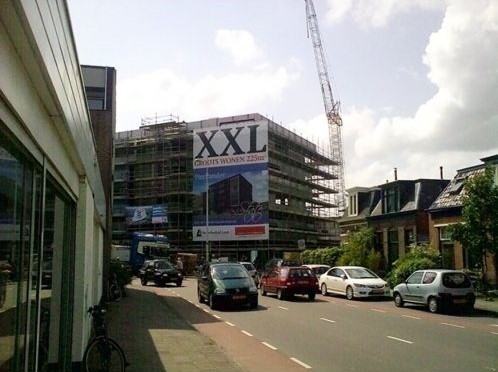 Gerüstbanner mit der Aufschrift 'XXL' an einem Gebäude im Bau, umgeben von Autos und einer Baustelle.