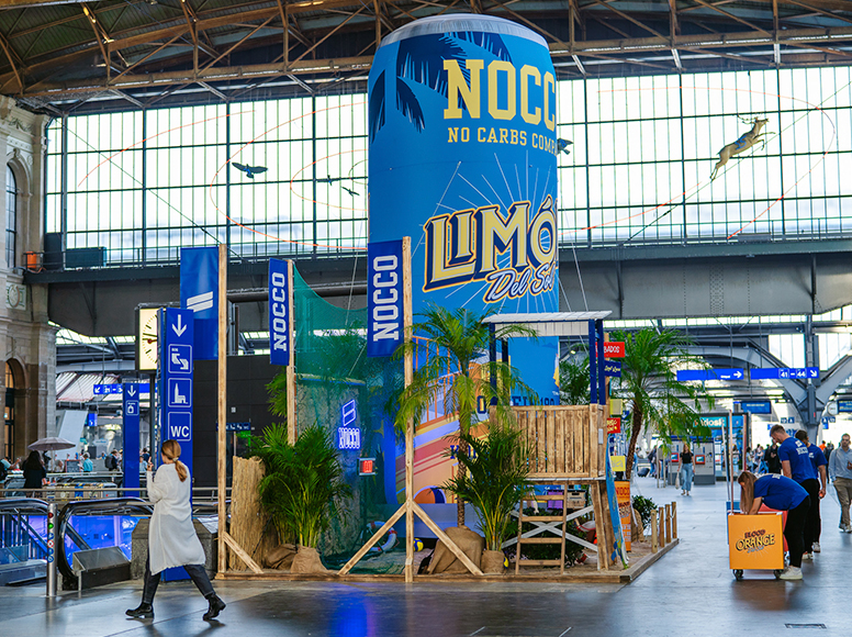 Aufblasbare Werbung für NOCCO Limonade in einem Bahnhof, umgeben von Pflanzen und Holzdekoration.