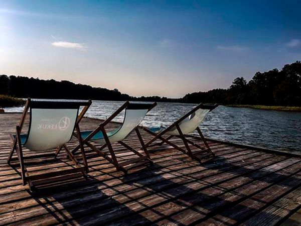 Drei Liegestühle aus Holz mit transparenten Stoffen auf einem Holzsteg am Wasser, umgeben von Bäumen.