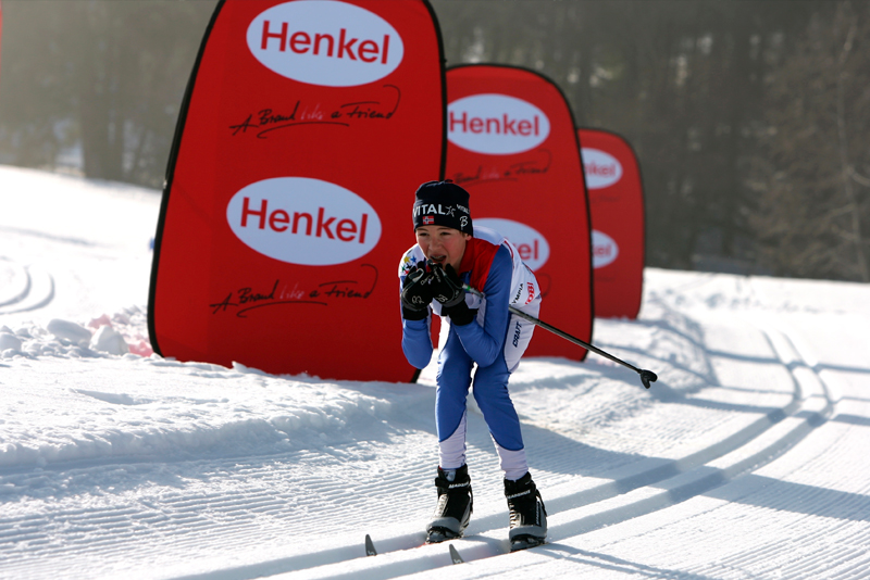 Skilangläufer in blauer Kleidung mit Skiern, der auf einer Loipe fährt, vor roten Werbebanden mit dem Logo von Henkel.