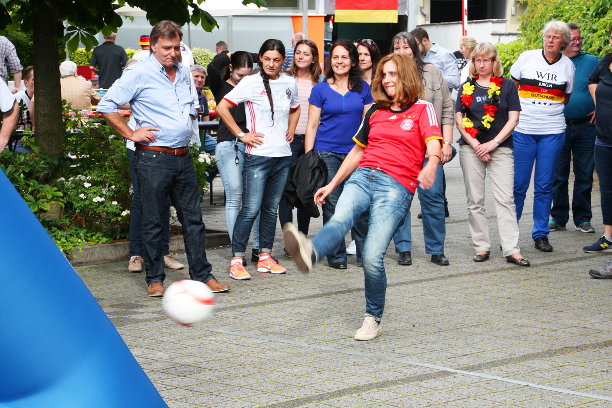 Ein Spieler in einem roten Trikot schießt auf eine Torwand beim Fußball-Kicker-Event. Zuschauer beobachten das Geschehen.