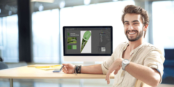 Ein lächelnder Mann sitzt an einem Tisch vor einem Computer, auf dem ein Layoutdesign angezeigt wird. Der Mann trägt ein helles Hemd und eine Armbanduhr.