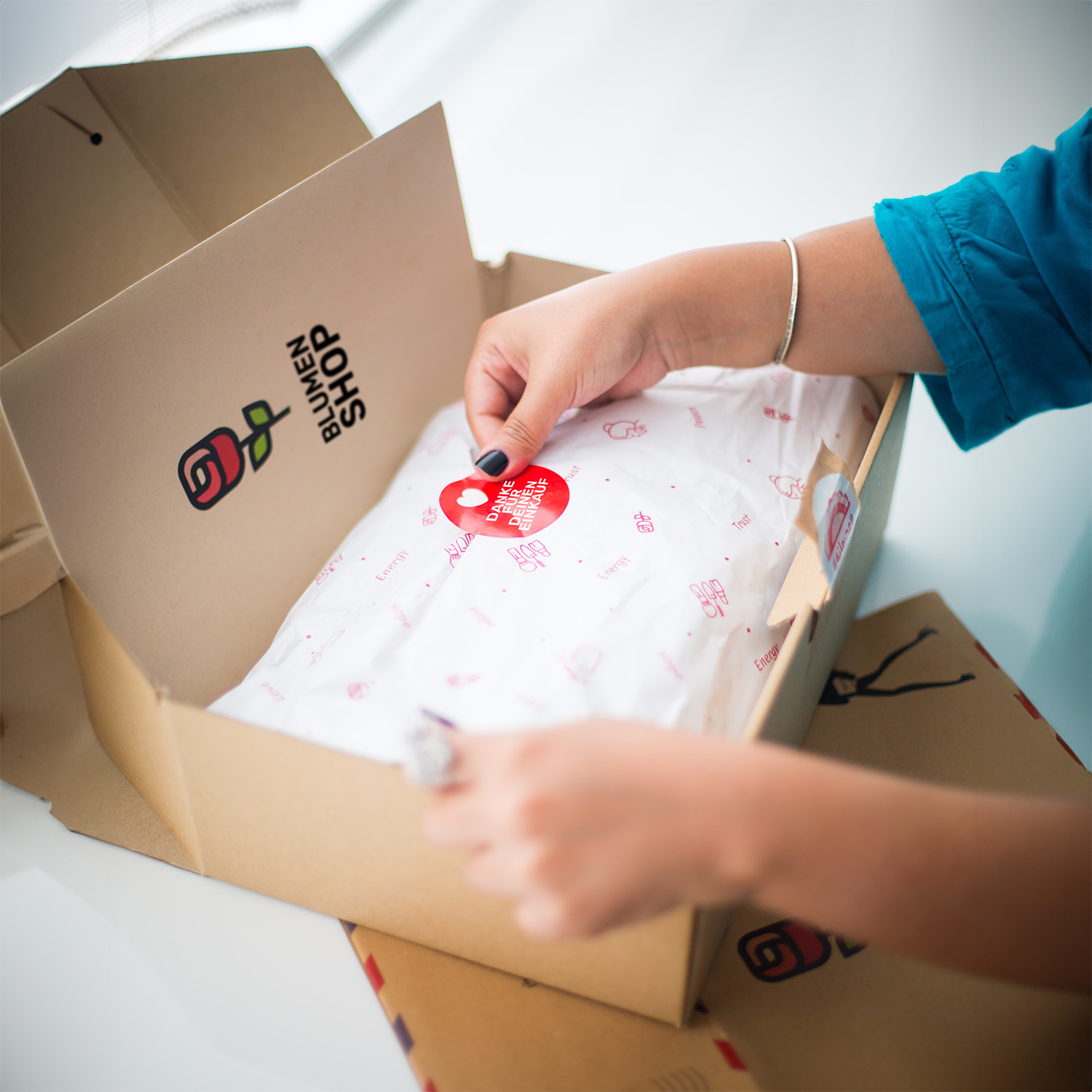 Eine Person öffnet ein Paket mit dem Aufdruck 'Blumen Shop', das mit rotem Papier und einem Aufkleber versehen ist.