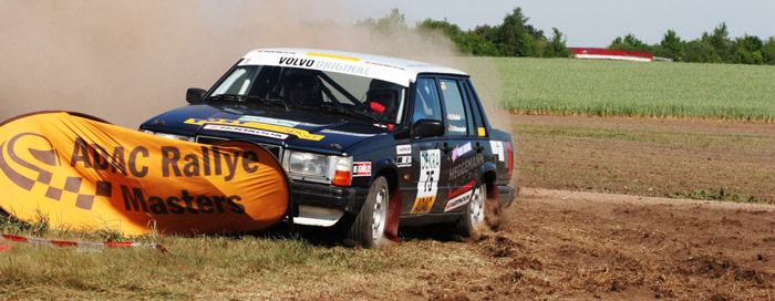 Ein Rallye-Auto fährt durch einen staubigen Feldweg und berührt eine Werbebande mit dem Aufdruck 'ADAC Rallye Masters'.