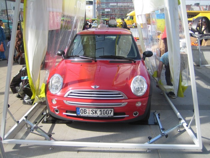 Roter Mini Cooper in einer Autotasche, umgeben von transparenten Wänden, auf einem Veranstaltungsgelände.