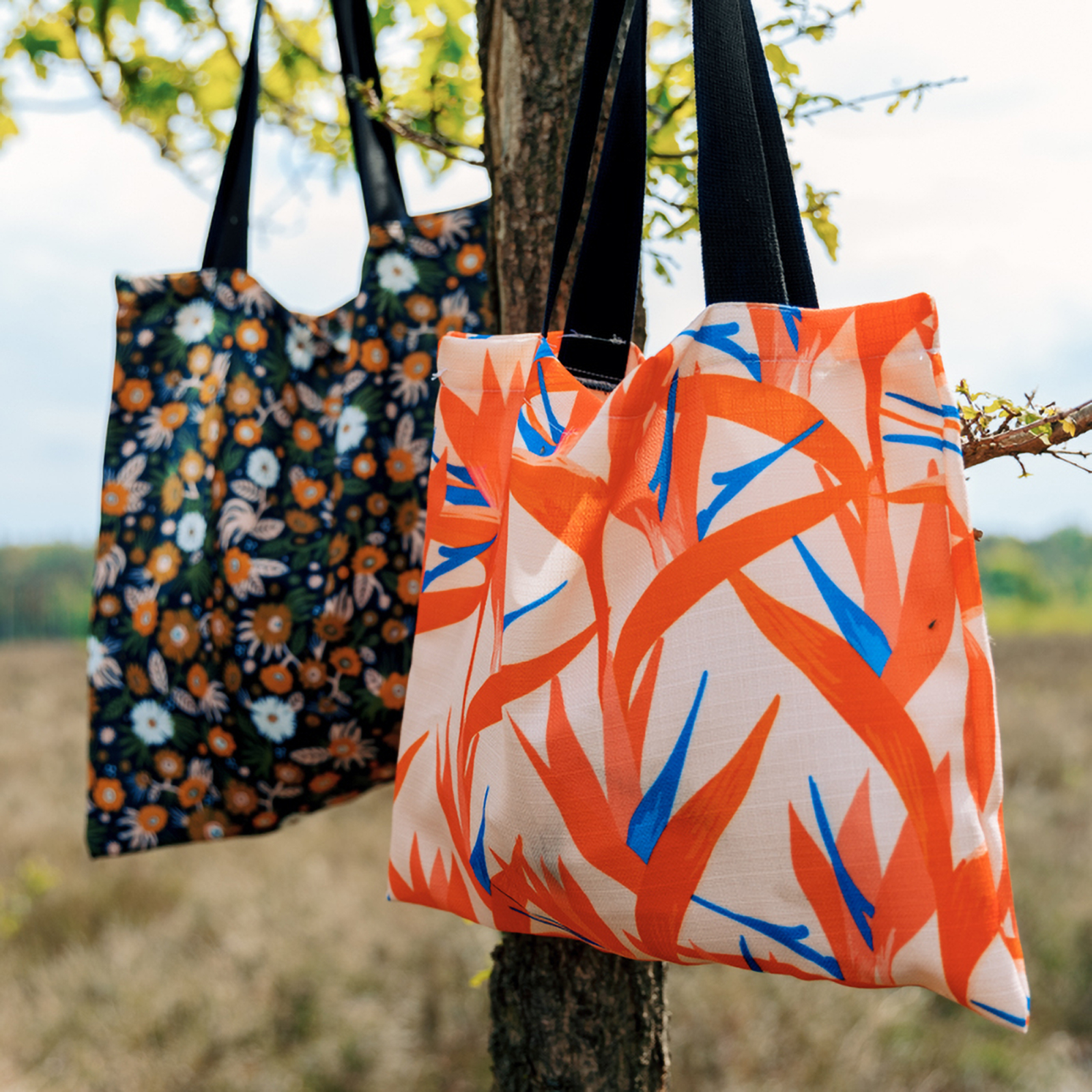 Zwei Tragetaschen von ENTDECKER mit bunten Designs, eine mit floralen Mustern und die andere mit abstrakten, orange-blauen Pflanzenmotiven, hängen an einem Baum.