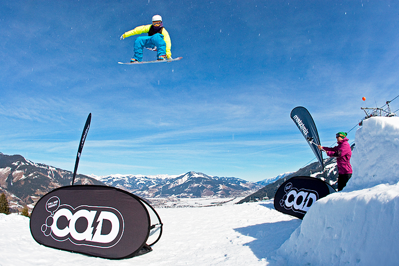 Snowboarder springt über eine Werbebande "Quickboard Winter" von ENTDECKER, während eine Zuschauerin zuschaut. Winterlandschaft im Hintergrund.
