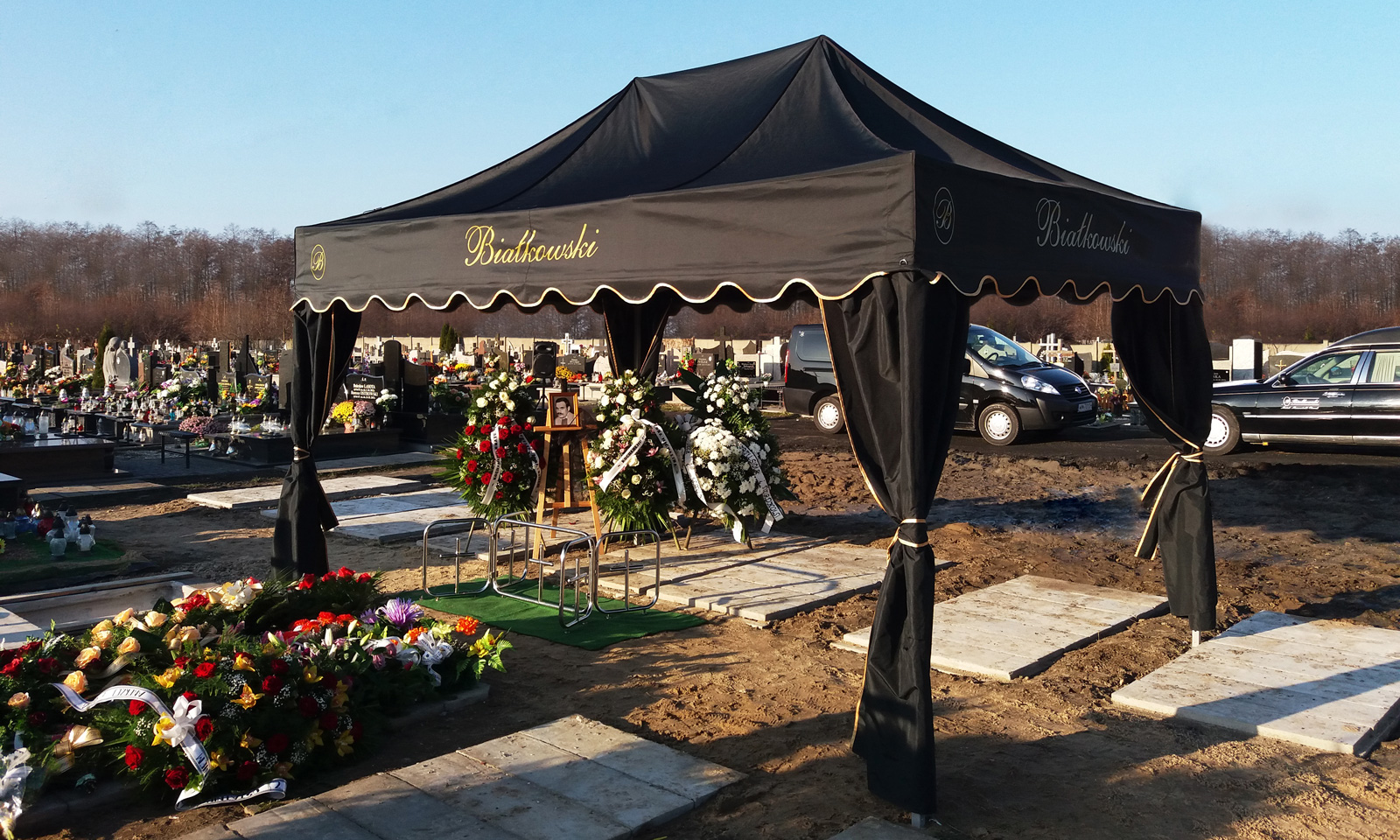 Schwarzes Beerdigungszelt mit goldener Schrift, umgeben von Blumenarrangements auf einem Friedhof.