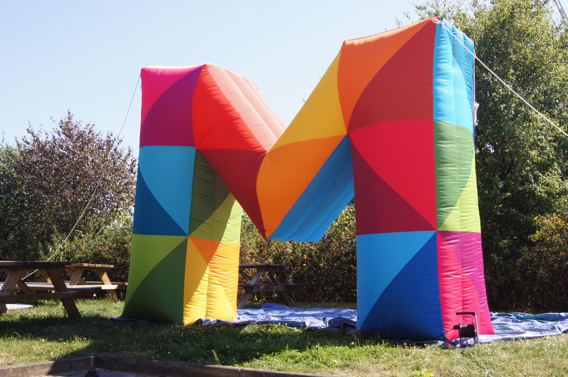 Buntes, aufblasbares M in verschiedenen Farben, auf einer Wiese mit Bänken im Hintergrund.