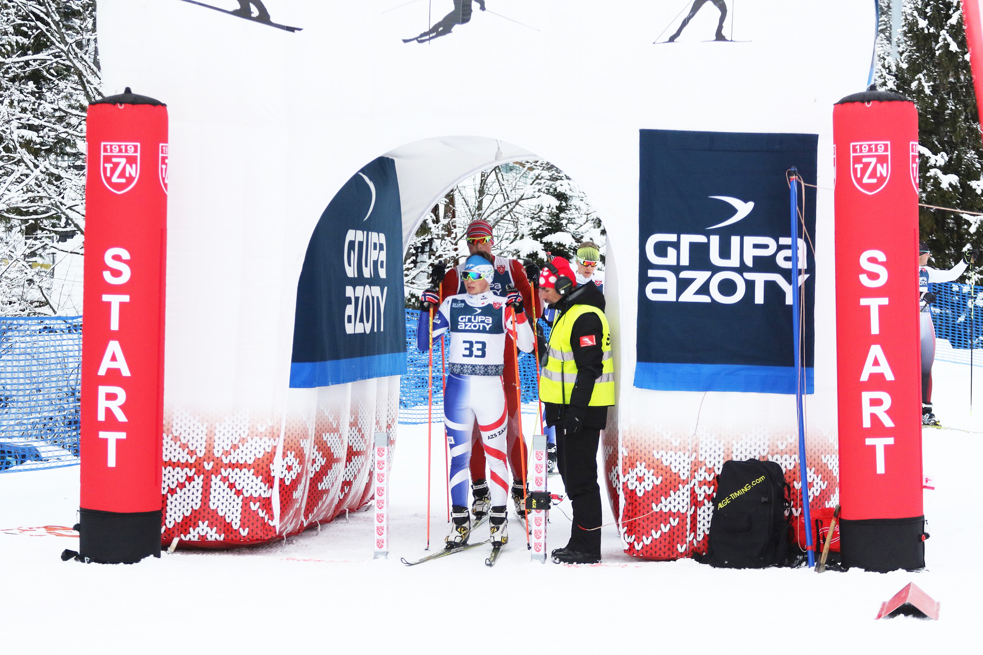 Drei Skifahrerinnen in roten Anzügen stehen an einer aufblasbaren Werbesäule von PZN während eines Wintersport-Events.