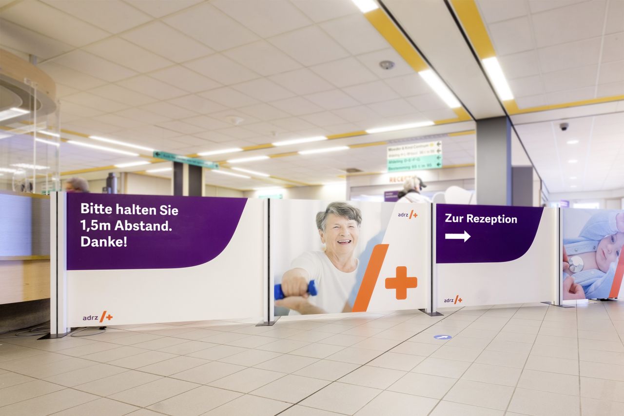 Absperrung mit dem Hinweis 'Bitte halten Sie 1,5m Abstand. Danke!' und einem Bild einer lächelnden Frau, platziert in einem Empfangsbereich.