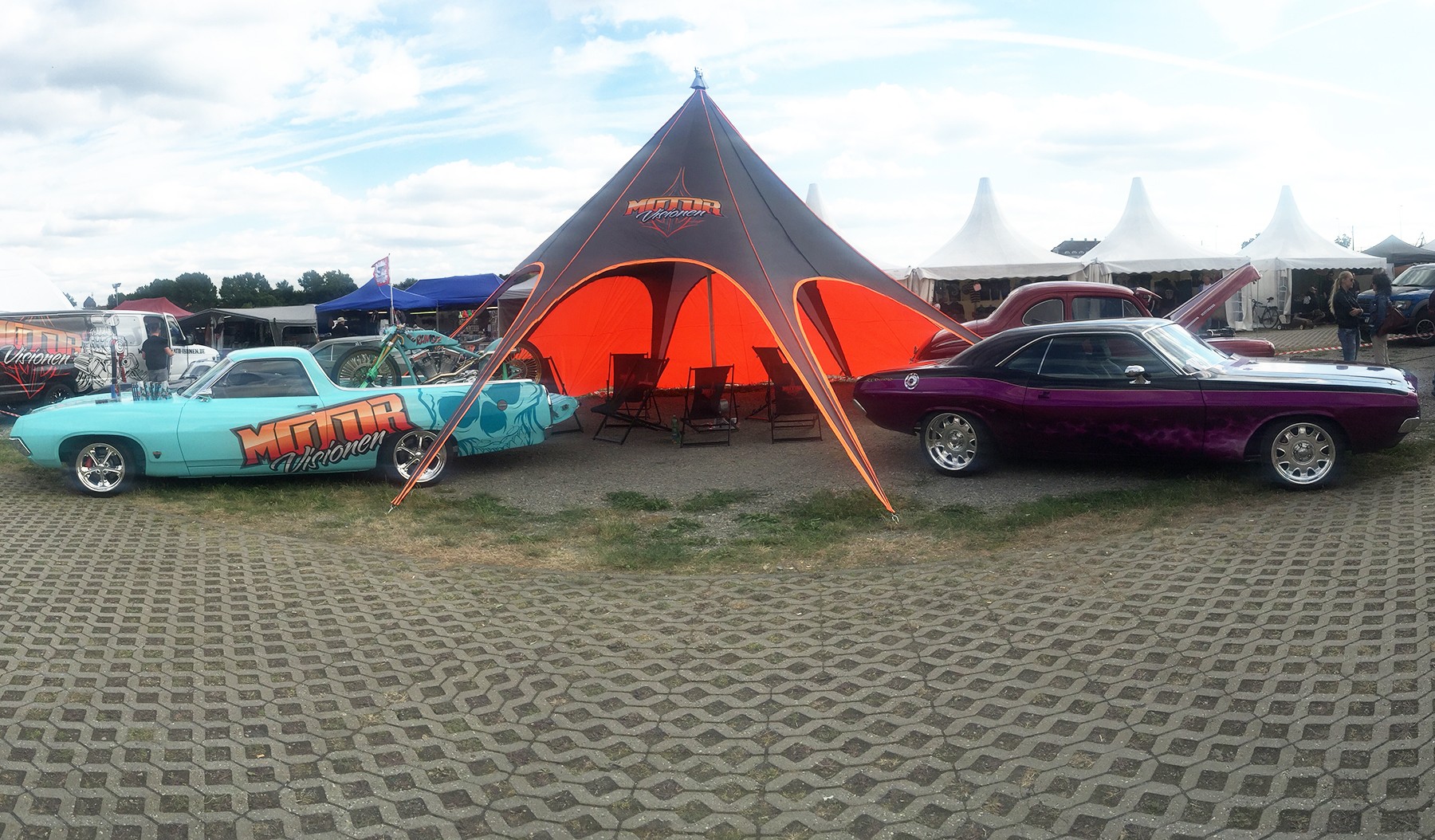 Sternzelt Jh Static Base von Motor Visionen, orange und schwarz, mit zwei klassischen Autos, einem blauen und einem lila Fahrzeug, auf einem Messegelände.
