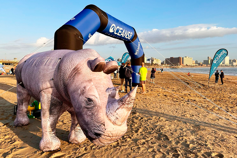 Aufblasbares Nashorn 'Nashorn Blow Up' steht am Strand unter einem Bogen mit der Aufschrift 'OCEANSB'.