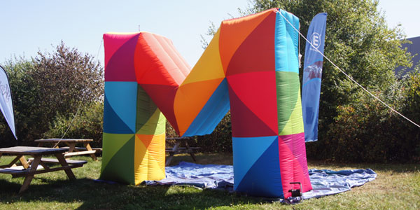 Bunte, aufblasbare M-Form in verschiedenen Farben auf einer Wiese, umgeben von Bäumen und einer blauen Plane.