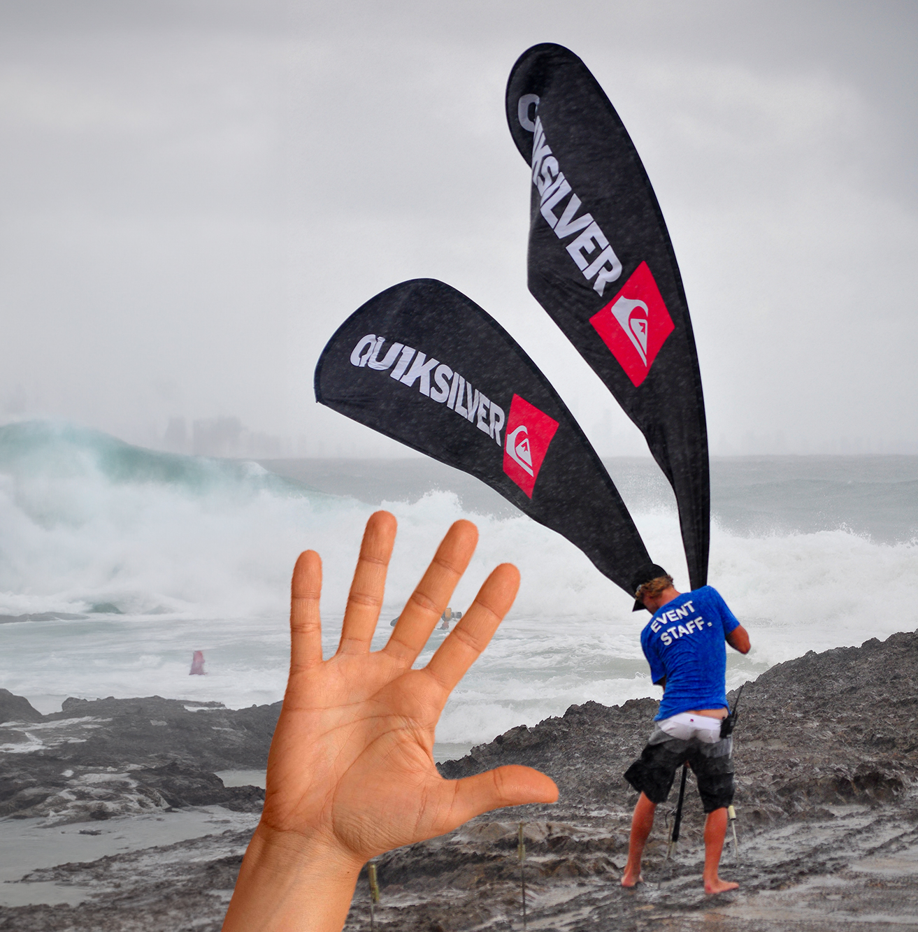 Ein Mitarbeiter in einem blauen T-Shirt mit der Aufschrift 'EVENT STAFF' hält zwei schwarze Fahnen mit dem Logo von Quiksilver am Strand bei stürmischem Wetter.
