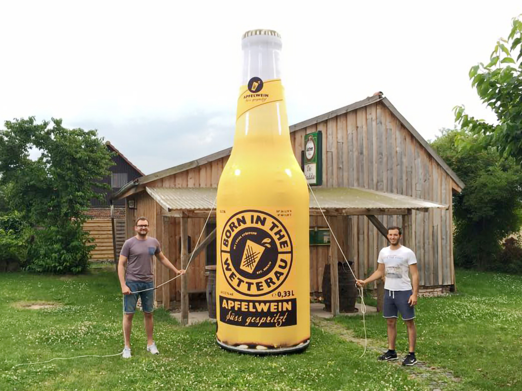 Zwei Männer halten eine große aufblasbare Flasche des Produkts 'Born In The Wetterau' mit der Aufschrift 'Apfelwein' im Freien.