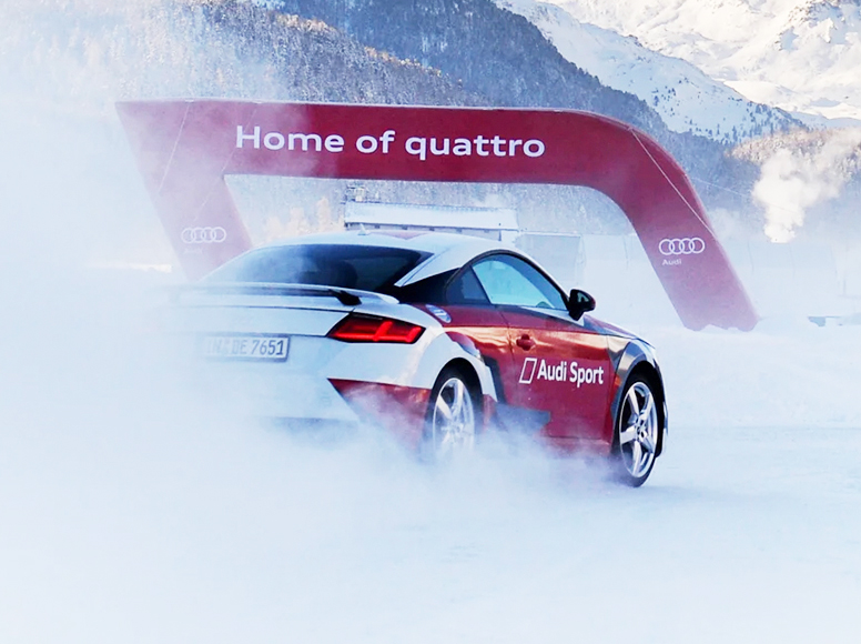 Audi TT Sportwagen auf verschneiter Strecke, hinter ihm ein aufblasbares Werbebanner mit der Aufschrift 'Home of quattro'.