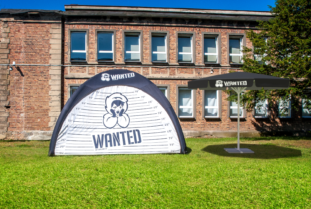 Zelt mit 'WANTED'-Aufdruck und Sonnenschirm auf einer Wiese vor einem Backsteingebäude.