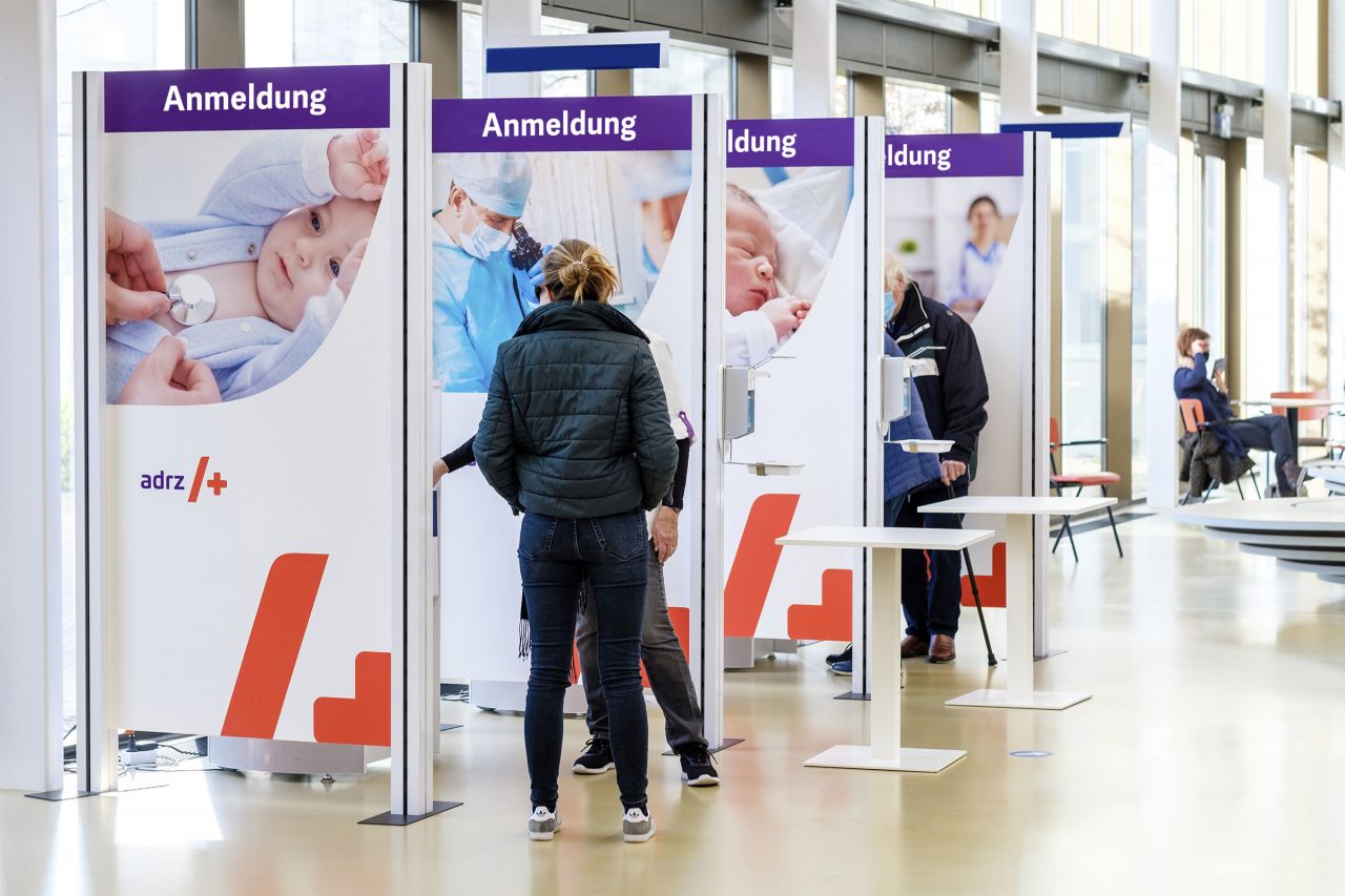 Raumteiler mit Anmeldeständen bei einer Veranstaltung, Personen stehen in Warteschlange.